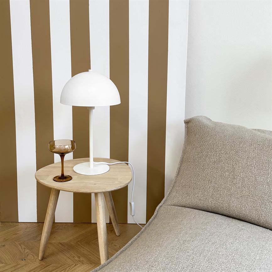 Et lille rundt træsidebord holder en hvid bordlampe og et brunt vinglas foran lodrette hvide og brune striber, ved en beige sofa på trægulv.