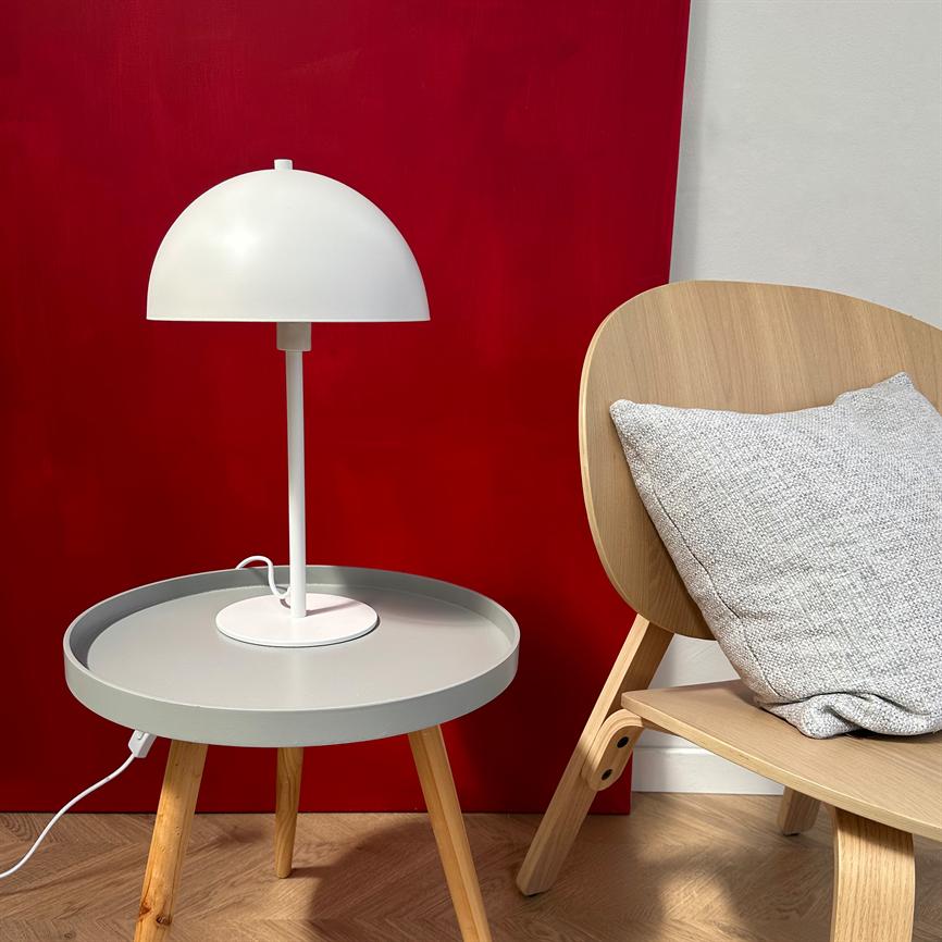 Hvid skærmlampe står på et rundt gråt sidebord med træben; foran en dyb rød væg, ved siden af en lys træstol med gråt pyntepudebetræk.
