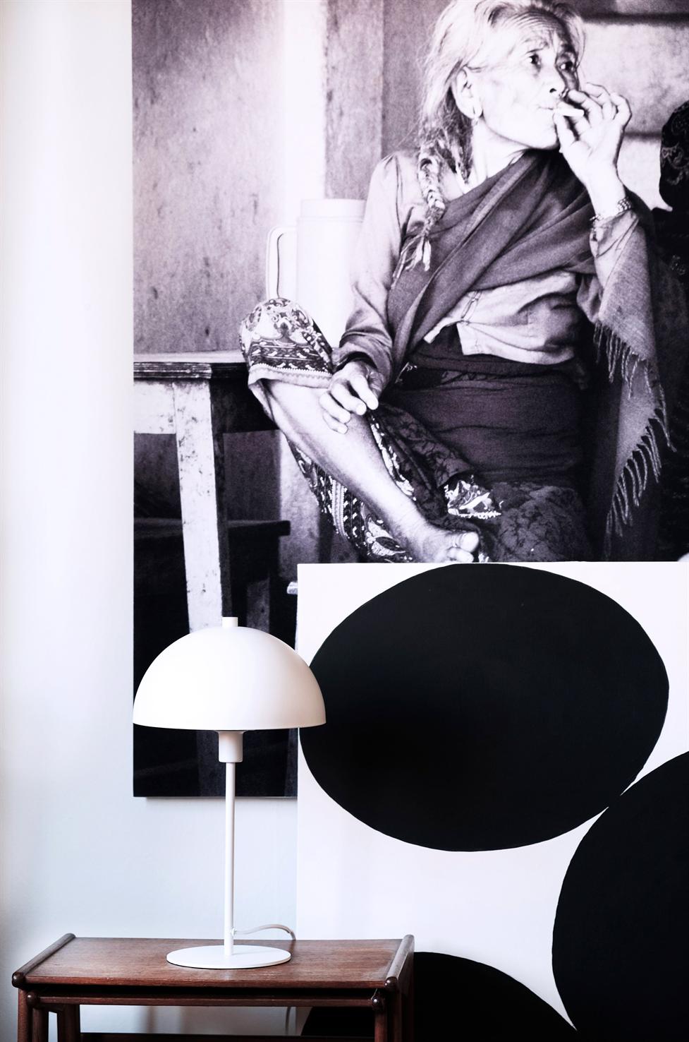 Lampe står på et træbord; foran et stort, grafisk lærred med sorte cirkler og et sort-hvidt foto af en ældre kvinde, der ryger, i baggrunden.