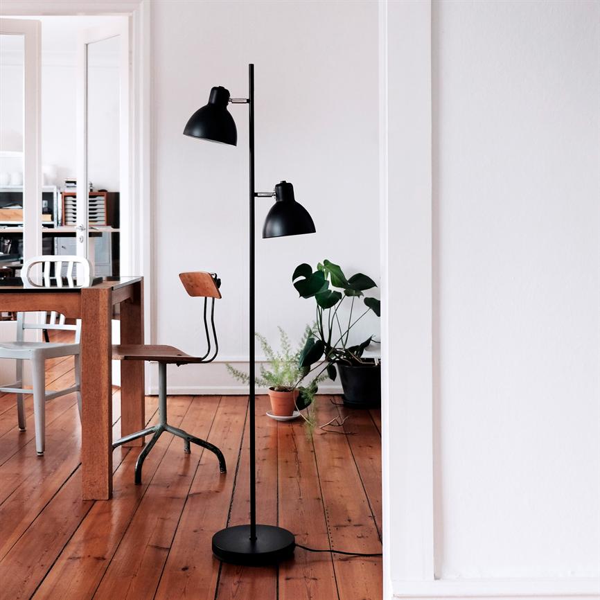 En sort gulvlampe står oprejst ved et træbord og en metal-træstol; i baggrunden potteplanter, hvide vægge og varme trægulvplanker i et lyst, minimalistisk spisestue-/arbejdsområde.