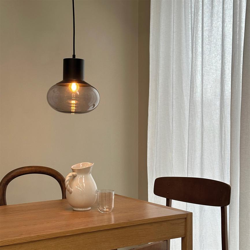 Hængelampe lyser med varm glød over et træbord; på bordet står en hvid kande og et glas; scenen omgives af to træstole og lyse, gennemsigtige gardiner.