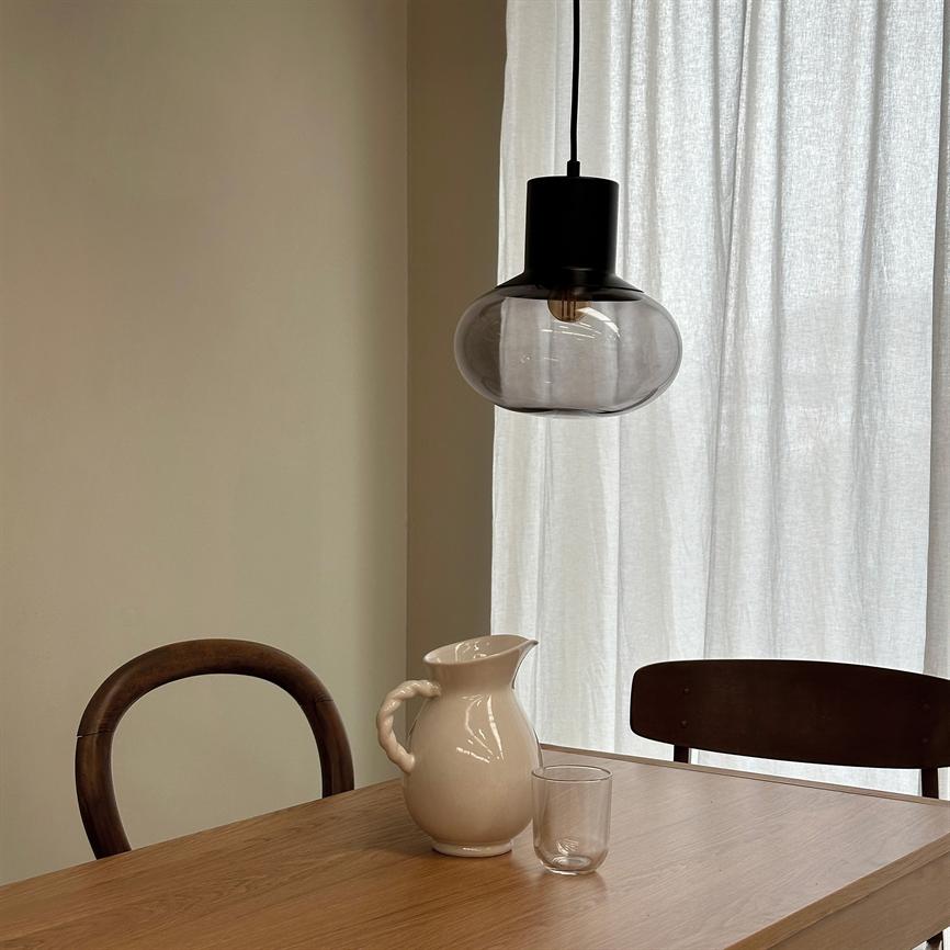 Hvid keramisk kande står på et træbord ved et klart glas; over dem hænger en sort pendellampe med røgfarvet glas, omgivet af to mørke træstole og et lysfilterende gardin.