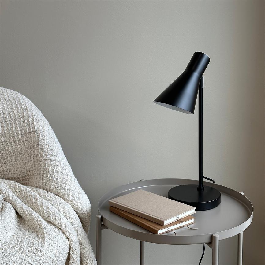 Sort bordlampe peger ned mod et rundt, gråt sidebord; to notesbøger ligger under lampen, og et cremefarvet, tekstureret tæppe er kastet over en nærliggende stol mod en neutral væg.