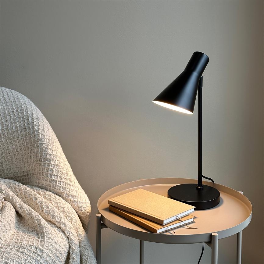 Sort bordlampe lyser ned på et rundt beige metalbord, hvor to notesbøger ligger stablet; ved siden af står et lyst, struktureret tæppe over en stol mod en neutral væg.