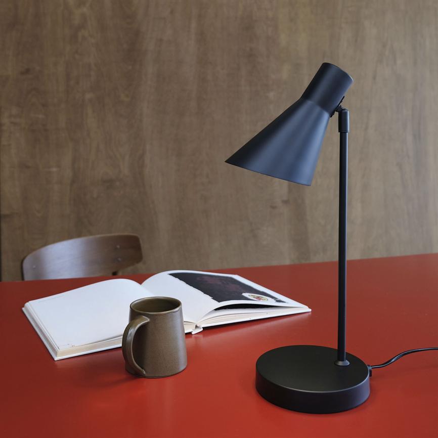 Sort bordlampe peger ned mod en åben bog og et brunt krus på et rødt bord, foran en træbeklædt væg og en træstol i baggrunden.