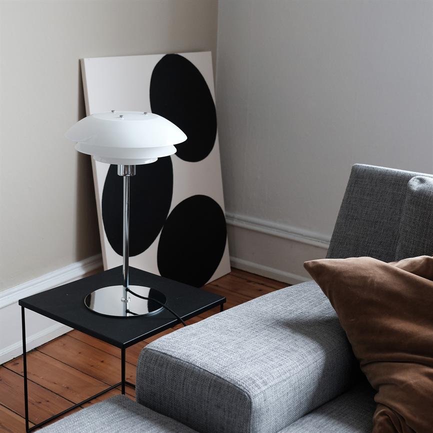 Bordlampe står slukket på et sort sidebord; ved siden af læner et hvidt maleri med tre store sorte cirkler mod en væg, foran en grå sofa på trægulv.
