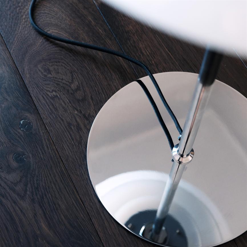Kromfod fra en gulvlampe står på mørkt trægulv; en blank, rund base spejler lampestammen, mens en sort, flettet ledning slynger sig henover basen og gulvet.