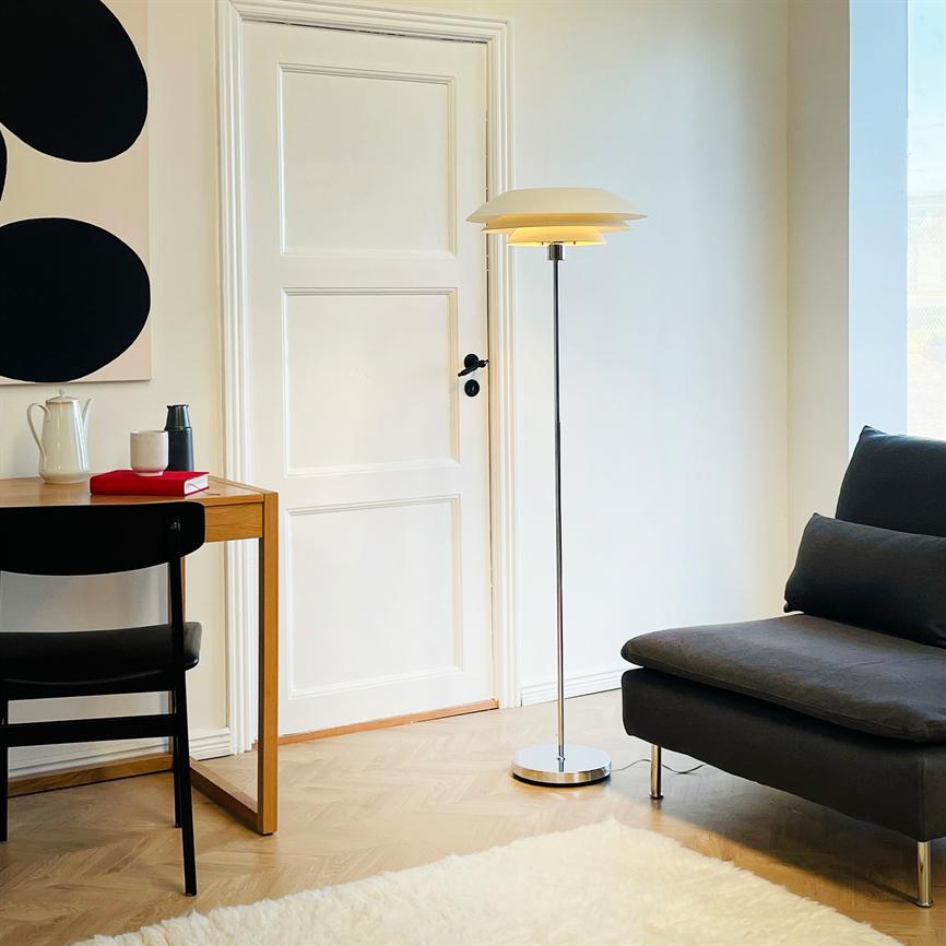 Gulvlampe står tændt og kaster blødt lys; i et lyst hjørne med en hvid dør og trægulv, ved siden af et træbord med sort stol og en mørkgrå sofa.