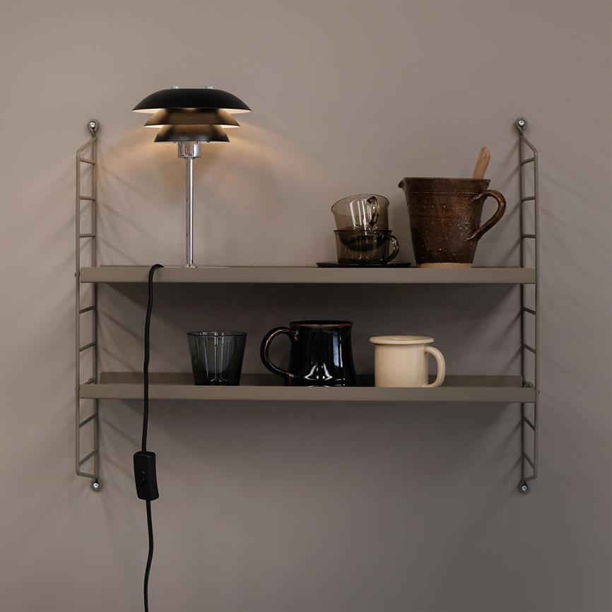En vægmonteret tohyldet metalhylde bærer en tændt bordlampe med sort skærm, glas- og keramikkopper, en rustik kande og en træske; ledning med tænd/sluk-knap hænger ned foran en beige væg.