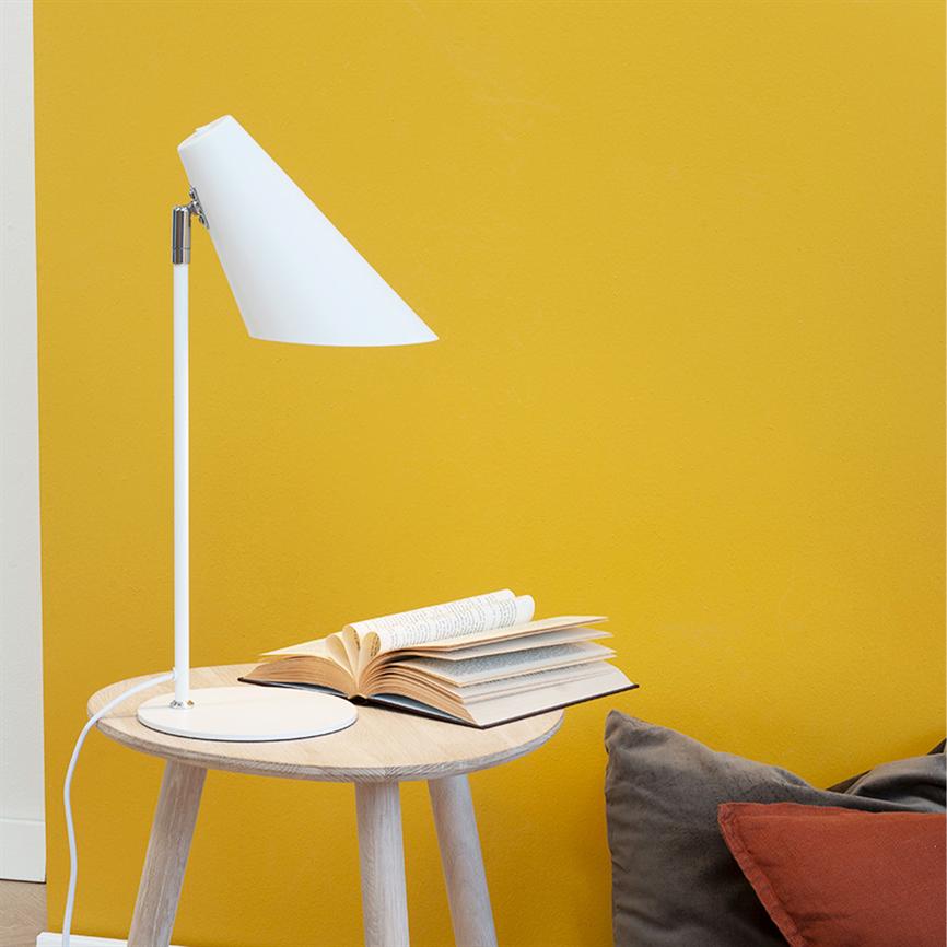 En hvid bordlampe vender sin skærm mod en åben bog på et rundt træbord, foran en varm gul væg med grå og brune puder ved siden.