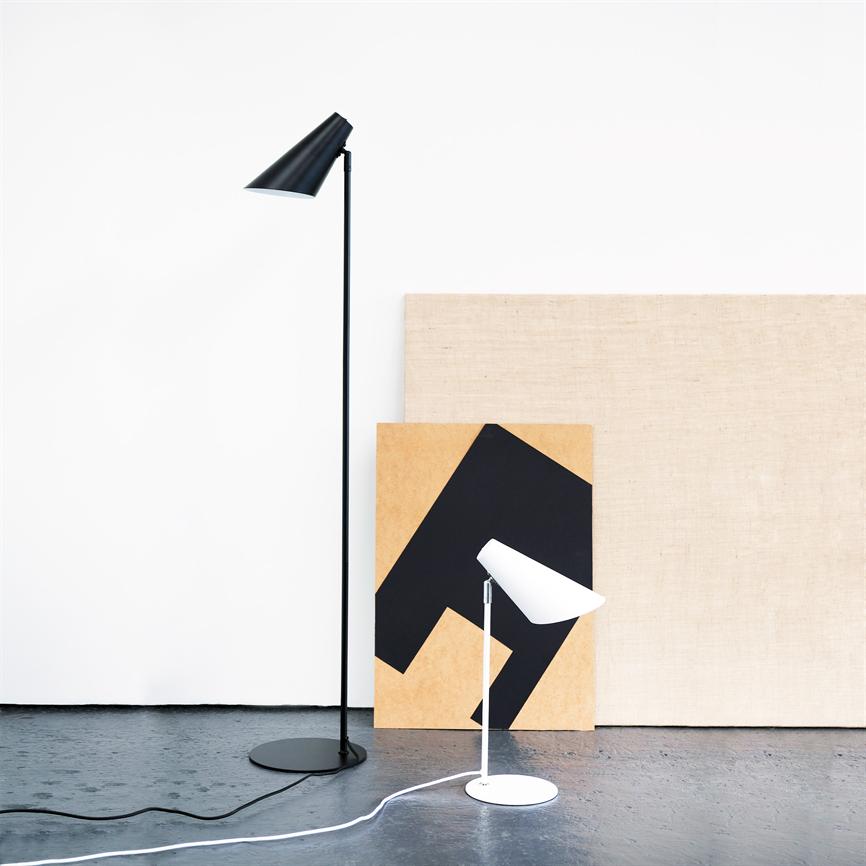 To lamper — en høj sort gulvlampe og en lav hvid bordlampe — står foran to lærreder og en hvid væg på et mørkt betongulv.