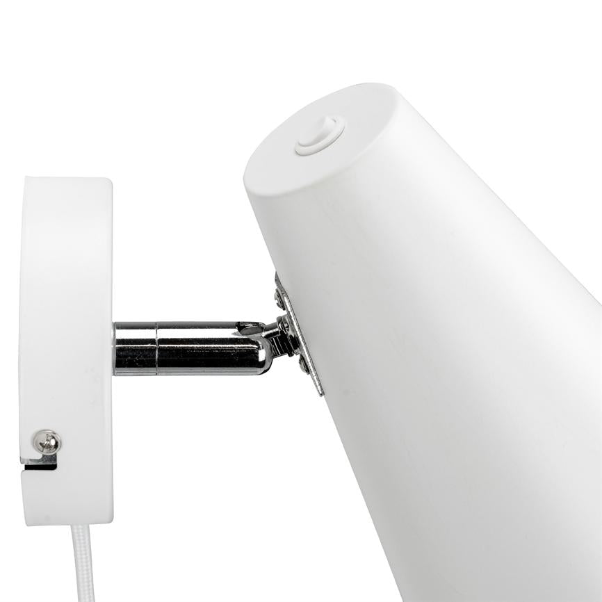 Hvid vægmonteret lampe peger skråt nedad, fastgjort med et kromled; hvid rund vægplade og synlig stofledning mod en ensartet hvid baggrund.