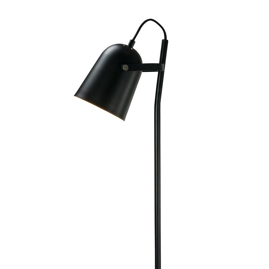 En sort metal gulvlampe hælder let fremad, lampeskærmen peger nedad og udsender blødt lys; slank stander mod en ensartet hvid baggrund.