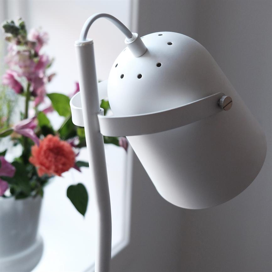 Hvid metallampe vipper skråt nedad, lampeskærm med ventilationshuller og slank stand; den står ved et vindue med en vase fuld af farverige blomster i blød fokus.