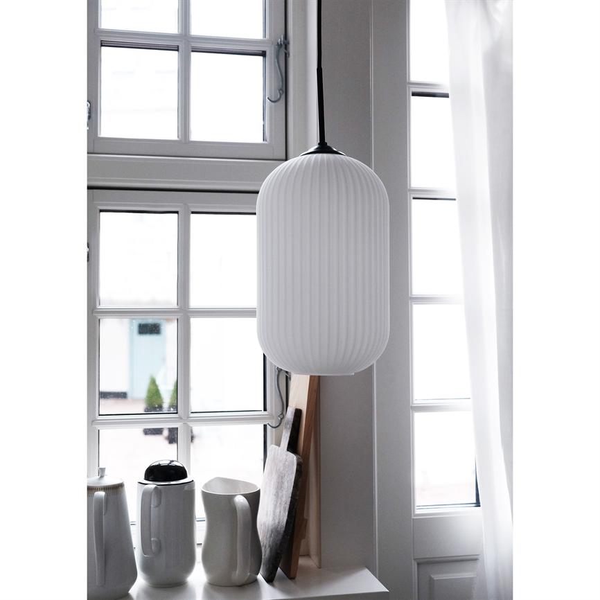 En hvid, ribbet pendel hænger foran et rutemasket vindue, oplyser en vindueskarm med hvide kander og skærebrætter i et lyst, minimalistisk køkken.