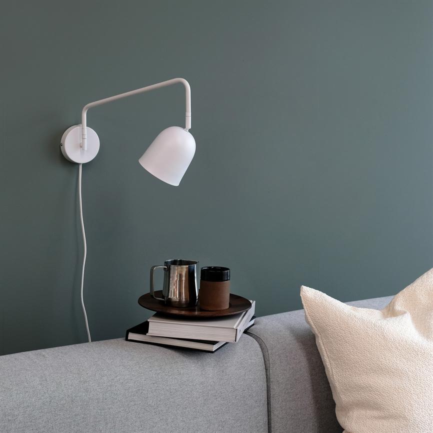 Hvid væglampe peger ned mod en bakke med bøger, et metalkrus og en termokop på armlænet af en grå sofa foran en grøn væg.