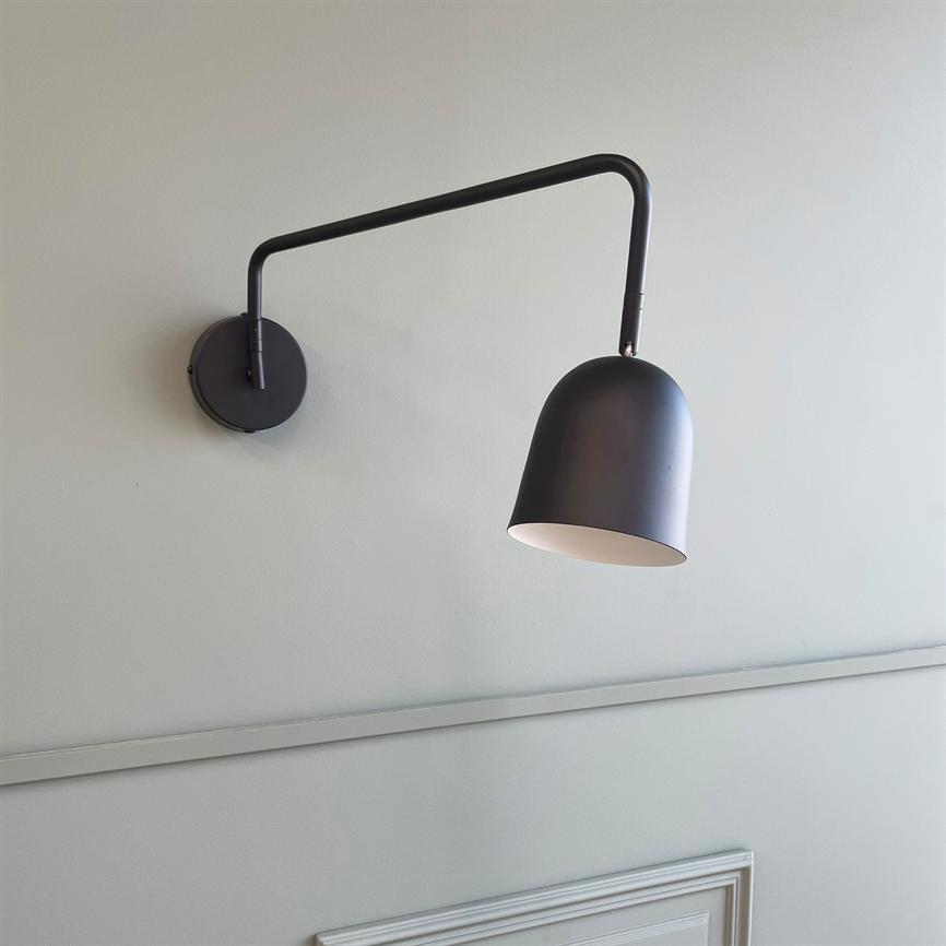 En sort vægmonteret lampe hænger på en lang buet arm og er vinklet nedad, med hvid inderside; den er monteret på en lys grå væg over en diskret panelliste.