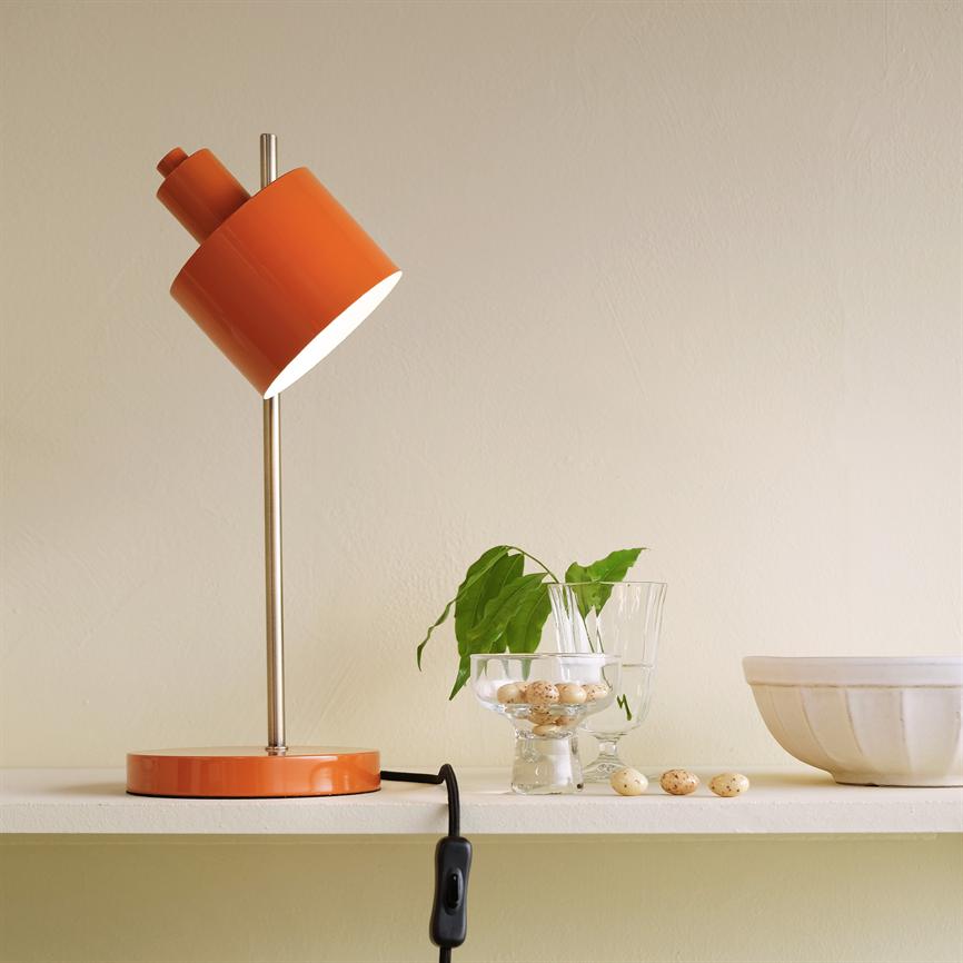 Orange bordlampe hælder og lyser på en hvid hylde ved en beige væg, flankeret af et glas med vagtelæg, en grøn stikling og en keramisk skål.
