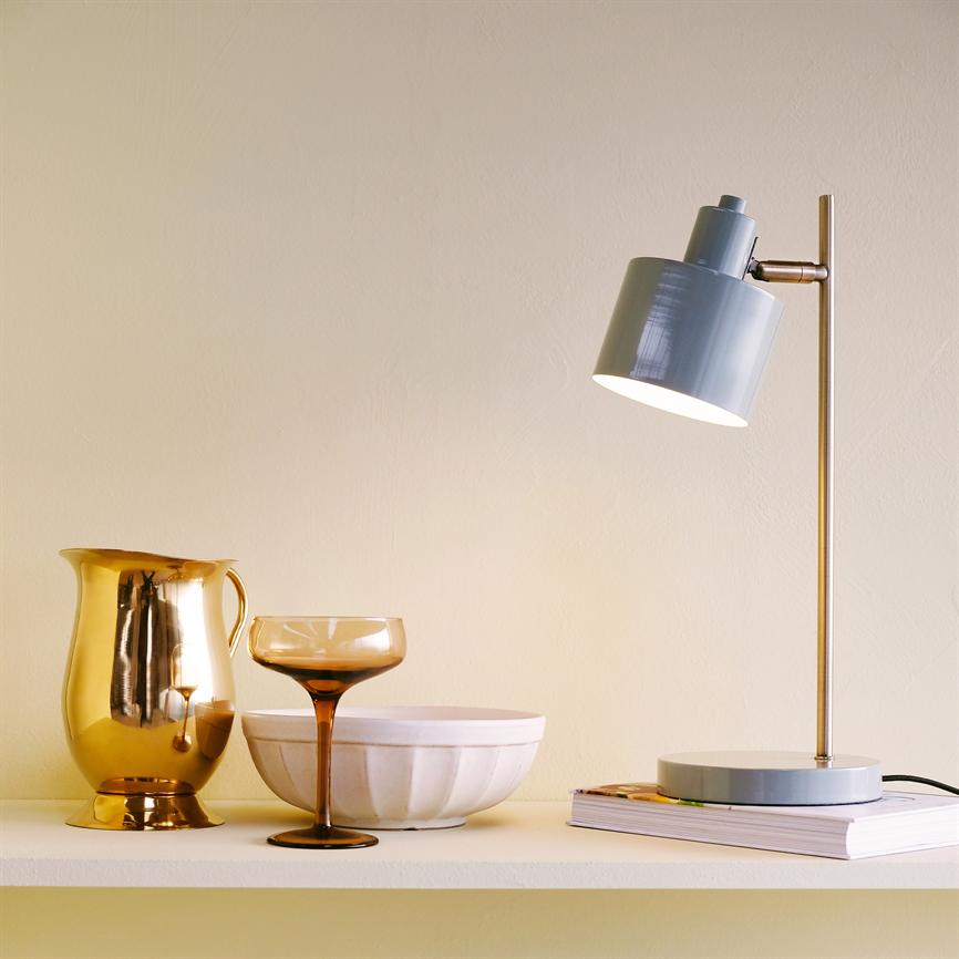 Bordlampe kaster blødt, varmt lys over en hylde med en gylden kande, et ravfarvet cocktailglas og en hvid skål; lampens metalfod står på en bog.