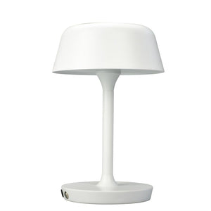 En hvid bordlampe står oprejst; skærmen buer let udad, monteret på en slank stang og rund fod; neutral hvid baggrund uden andre genstande.