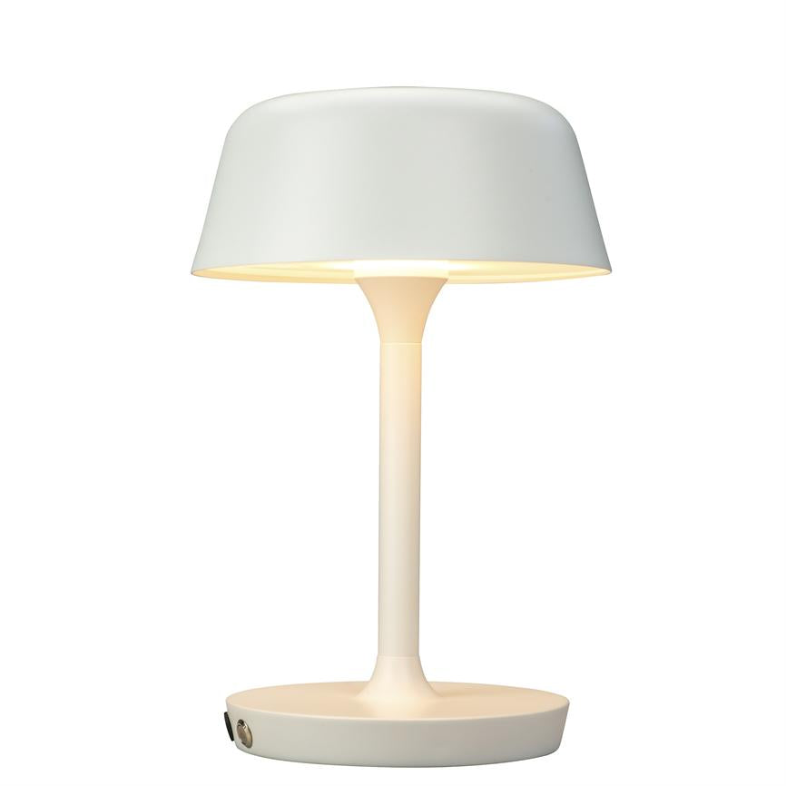 Bordlampe lyser blødt; slank cylindrisk stander, bred afrundet skærm og lav rund fod på neutral hvid baggrund.