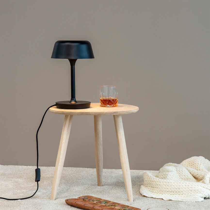 En sort bordlampe står på et rundt lyst træbord ved siden af et glas med mørk væske; kablet hænger ned, og et træbræt samt et strikket tæppe ligger på beige gulv.