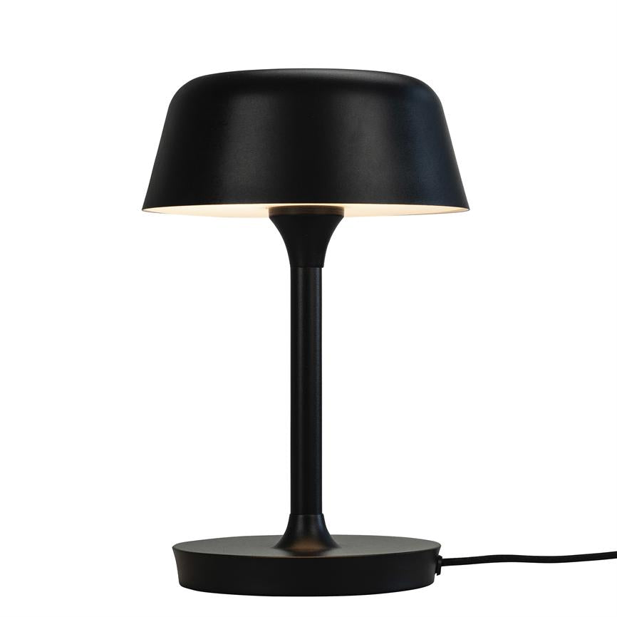 Bordlampe med sort rund fod og slank stang, udsender blødt nedadrettet lys gennem en bred sort skærm, mod hvid baggrund.