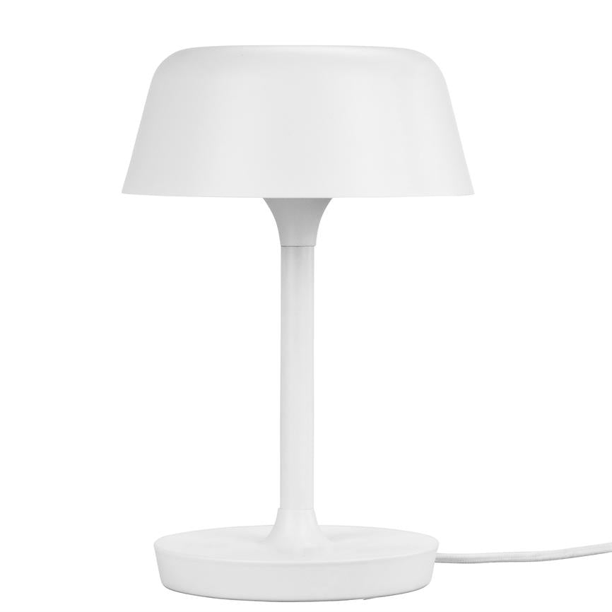 En hvid bordlampe står oprejst, med rund skærm og slank søjle, placeret på en flad rund fod; ledning løber ud til højre mod en hvid baggrund.