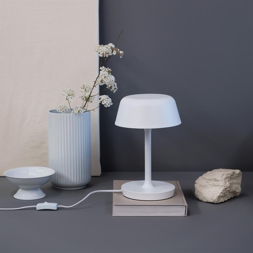 Hvid bordlampe står på en bog med synlig ledning, på en grå flade foran en mørkegrå væg; til venstre en rilleret vase med hvide blomster og en skål, til højre en rå sten.