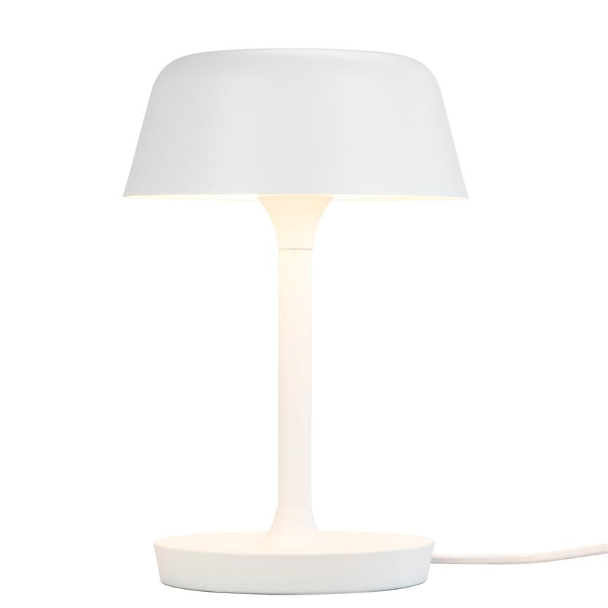 En hvid bordlampe står tændt; skærmen kaster blødt lys ned over en slank søjle og rund fod, omgivet af en ensartet hvid baggrund.