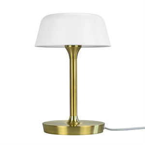 En bordlampe står med hvid skærm på en slank, børstet messingstang over en rund messingfod med synlig hvid ledning, isoleret mod hvid baggrund.