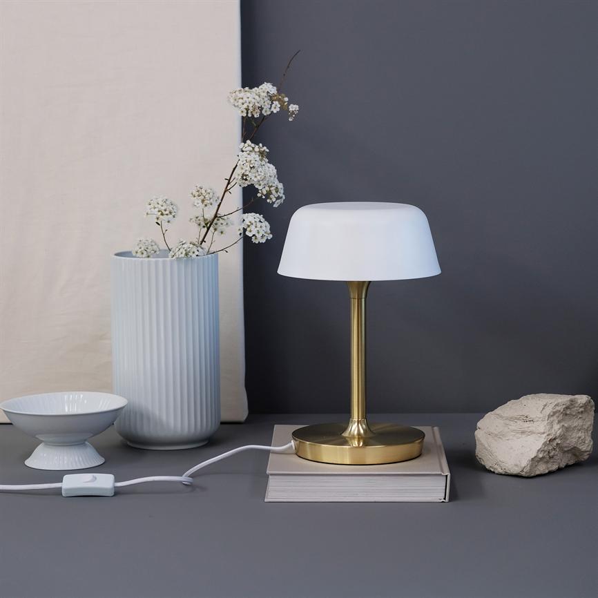 Bordlampe står på en bog, med hvid skærm og messingstamme; ved siden af en høj rillede vase med små hvide blomster, en skål og en rå sten på et gråt bord.