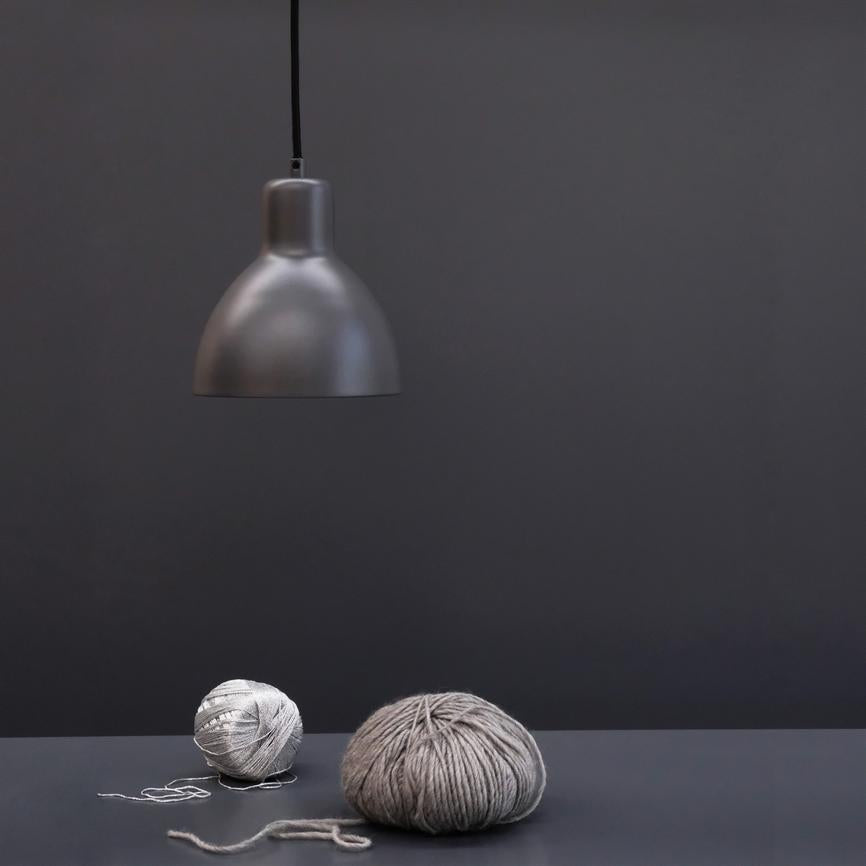 En mat grå loftslampe hænger over to grå garnnøgler, der ligger på et mørkegråt bord i et minimalistisk, ensfarvet gråt rum.