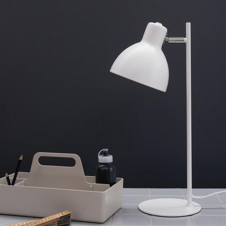 Hvid skrivebordslampe peger nedad og står slukket på en rund fod; foran en beige opbevaringsbakke med blyanter, flaske og lineal på flisebelagt bord mod en mørkegrå væg.