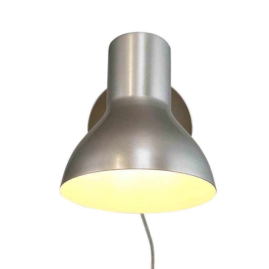 Sølvfarvet væglampe med rund skærm, lyser varmt nedad; monteret på et rundt vægbeslag, med et synligt kabel, fotograferet mod neutral hvid baggrund.