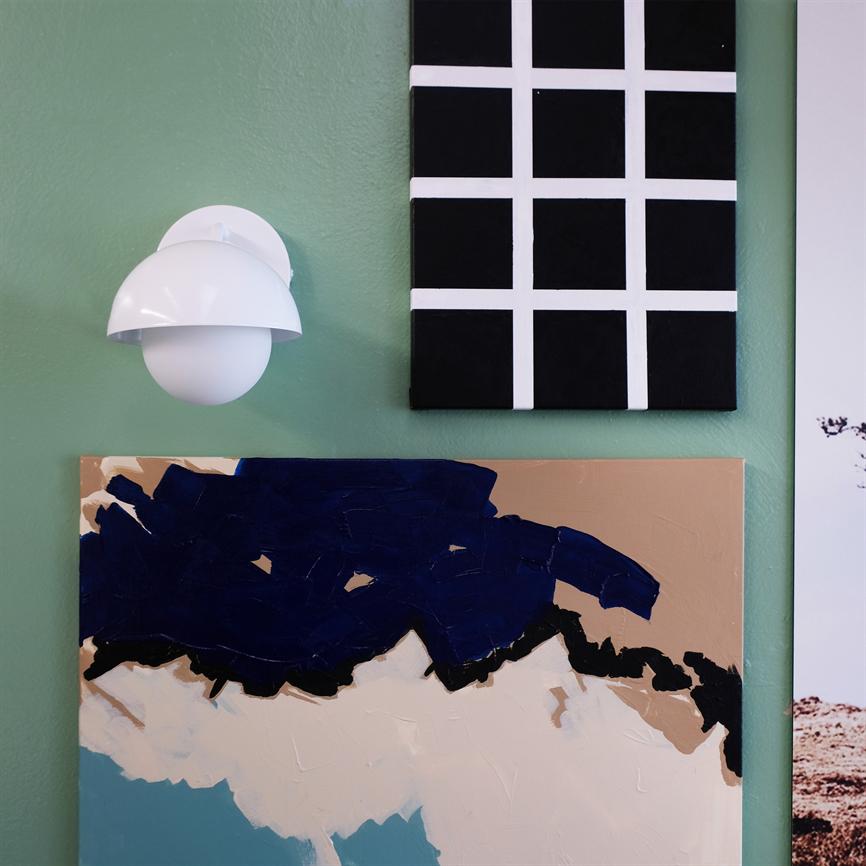 En hvid væglampe er monteret på en grøn væg, omgivet af to moderne malerier: et sort lærred med hvide ruder og et abstrakt lærred i mørkeblå, beige og turkis.