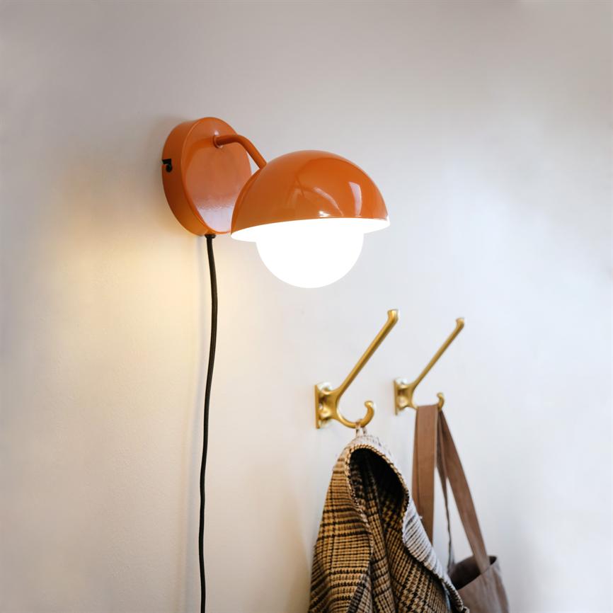 Orange væglampe lyser nedad, hænger fra sort ledning; under den sidder to messingkroge med en ternet frakke og en brun taske på en lys væg.