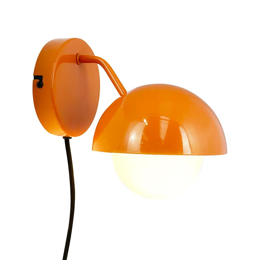 En orange væglampe hænger monteret, en arm fører til en halvskærm over en hvid rund opalglasdiffuser, som lyser svagt; sort stofledning hænger ned i en hvid baggrund.
