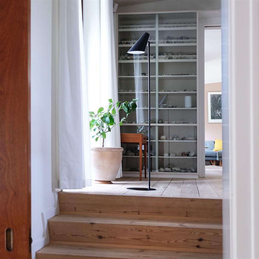 Gulvlampe står vippet mod gulvet ved en stor krukkeplante i et lyst rum med trætrappe, glasvitrineskab fyldt med små sten og en sofa i baggrunden.