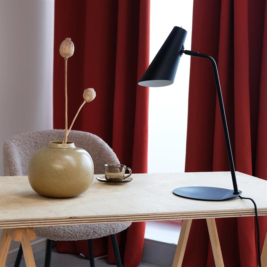 Sort skrivebordslampe, vinklet ned mod bordpladen; står på et lyst krydsfinerbord ved en beige vase med tørrede frøkapsler og en lille glas kop, foran dybrøde gardiner.