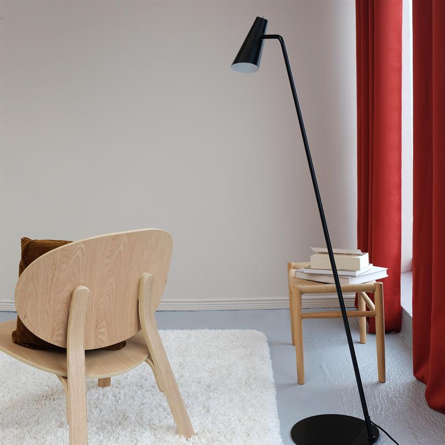 Sort gulvlampe bøjer sig over en lille træskammel med en stak bøger i et minimalistisk rum med lyst træstol, hvidt blødt tæppe og røde gardiner ved vinduet.