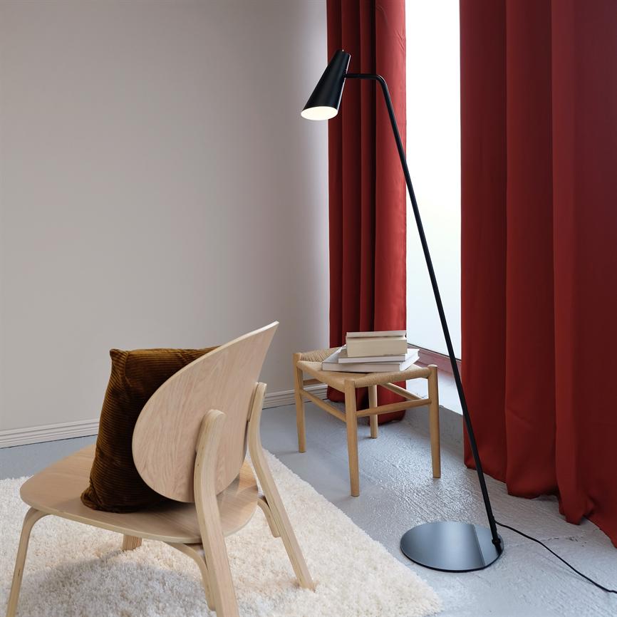 En slank sort gulvlampe kaster lys ned over et lyst træstol og et lille bord med bøger, ved et stort vindue med tunge røde gardiner i et minimalistisk, lyst rum.