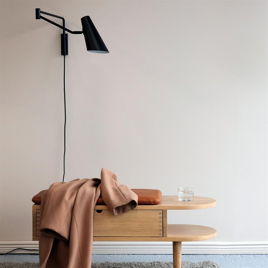 Et lille træbord med skuffe har en beige jakke draperet over sig og et glas vand; en sort væglampe er monteret over bordet mod en lys væg og blåt tæppe.