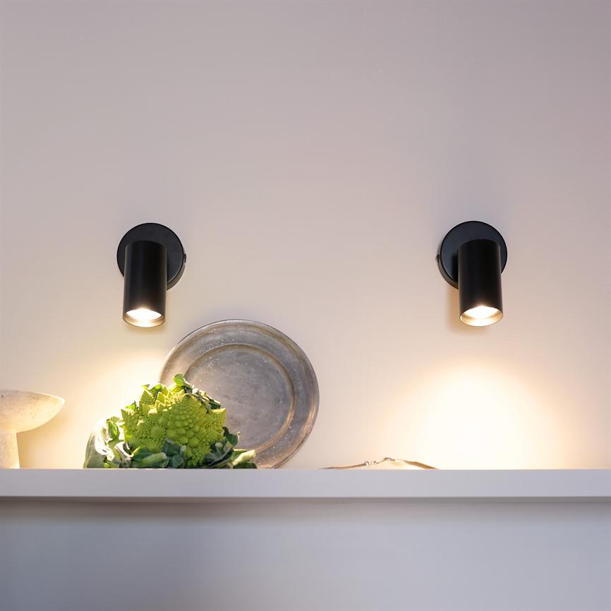 To sorte væglamper lyser nedad over en hvid hylde, hvor en romanesco-blomkål ligger foran et rundt metalfad mod en neutral, lyst malet væg.