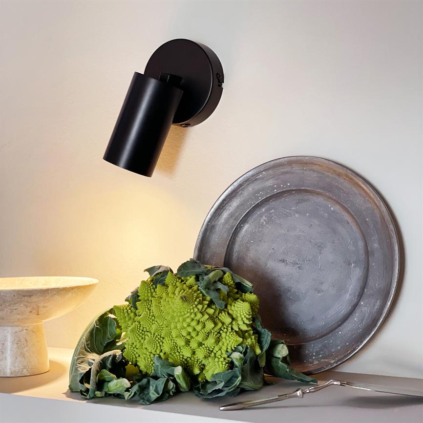 En grøn romanesco-broccoli ligger på en hylde under en sort væglampe, flankeret af et rustikt metalfad, en marmor-skål og et par sølvfarvede serviceværktøjer.