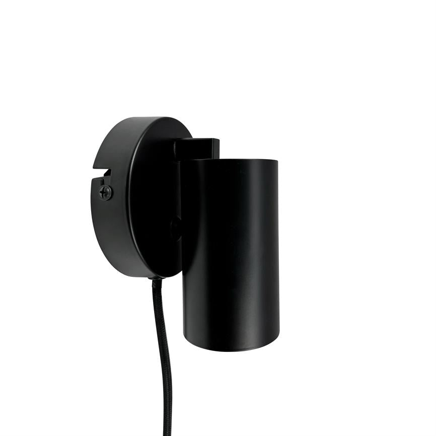 Sort cylindrisk væglampe monteret på rundt vægbeslag, med tekstilbeklædt ledning hængende ned; minimalistisk design mod en ren, hvid baggrund.