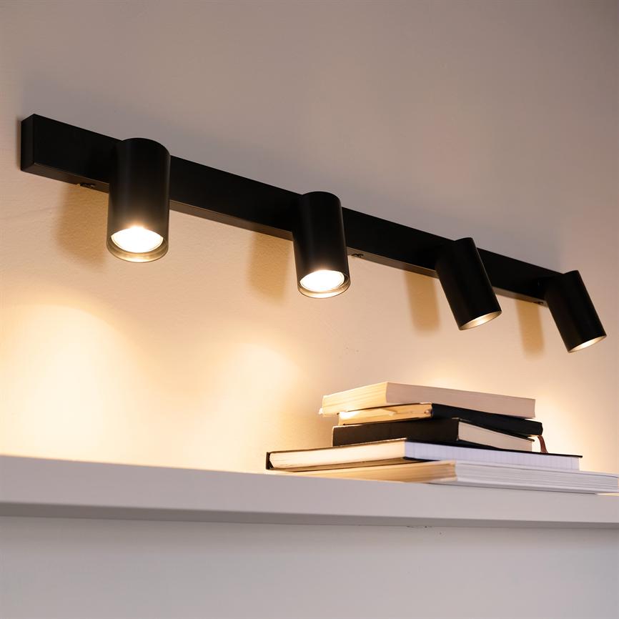 En sort, fire-armet spotlampe monteret på væggen lyser nedad og kaster varme, cirkulære lyspletter over en hvid hylde med en stak bøger.