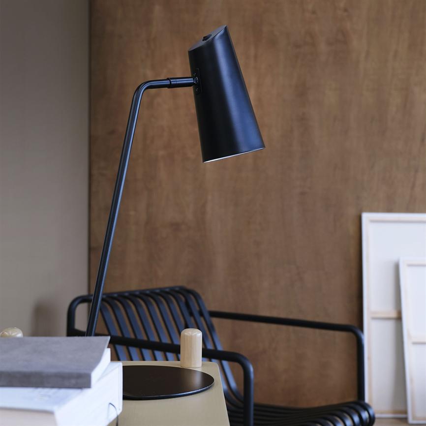 Sort skrivebordslampe peger nedad over et sidebord i et moderne interiør; omgivet af stablede bøger, en sort metalstol med lameller og en træbeklædt væg med opstående lærreder.