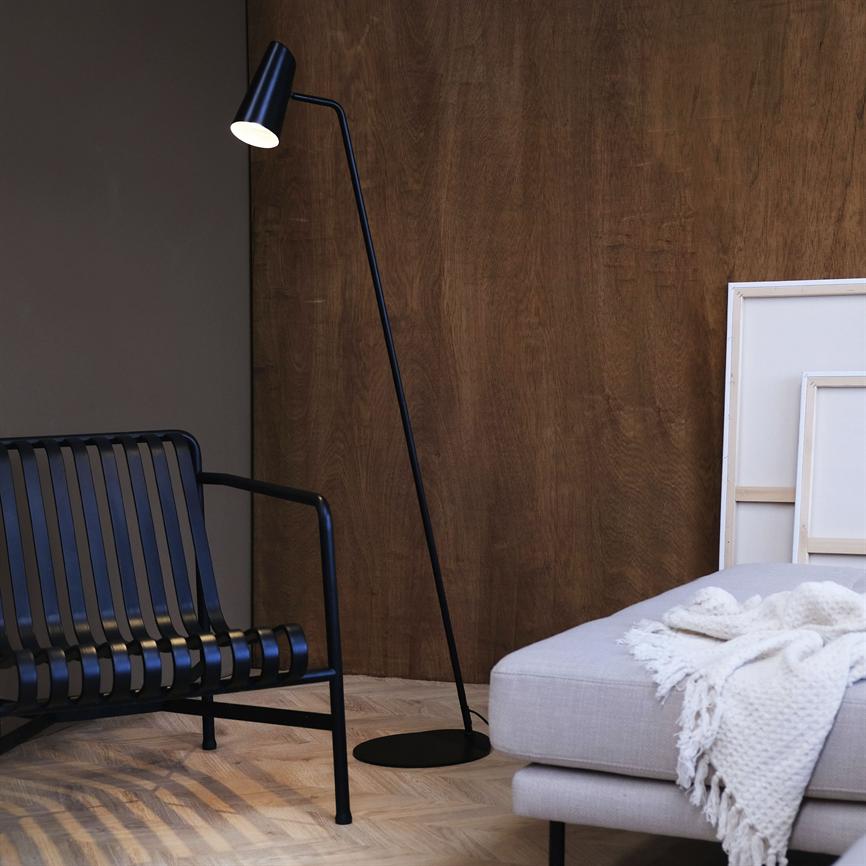 En sort, slank gulvlampe står tændt og kaster lys mod gulvet; i et moderne interiør med sort metalbænk til venstre, lysegrå sofa med strikket tæppe til højre og mørk trævæg.