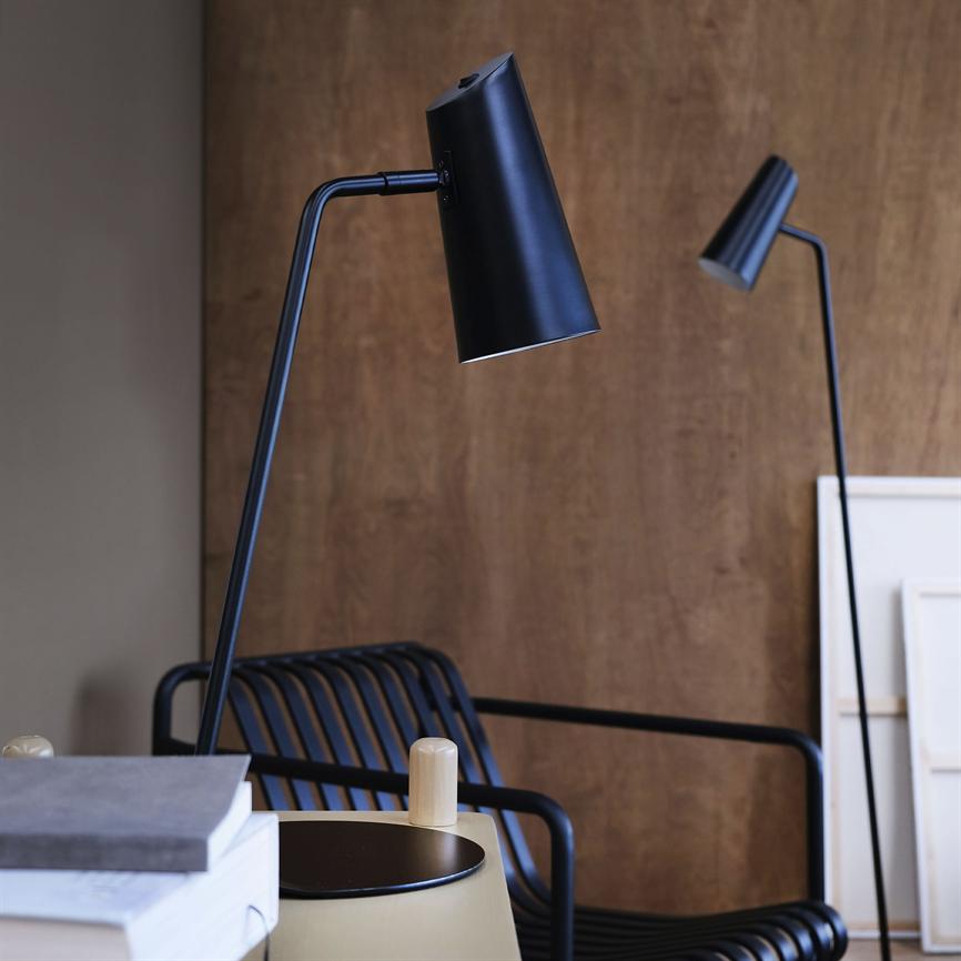 Sort kegleformet lampe bøjer fremad og lyser ned mod et lille rundt bord; foran en sort metalstol med lameller og en væg i træ, med bøger i forgrunden.